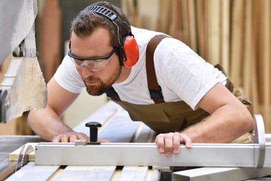 Tischler in einer Schreinerei sägt ein Holzbrett an einer Sägemaschine - Handwerker in Berufsbekleid
