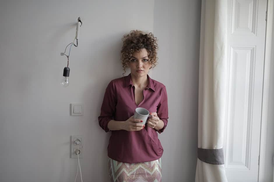 Gloomy female with curly hair in blouse and skirt leaning on wall in empty unfinished room while sta
