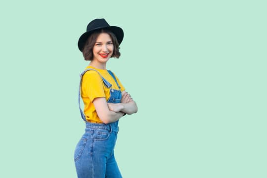 Happy pretty young hipster girl in blue denim overalls, yellow shirt and black hat standing, crossed